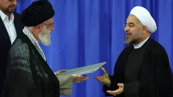 L'image montre deux hommes en tenue traditionnelle, se tenant debout l'un en face de l'autre. L'un d'eux porte un turban noir et une cape sombre, tandis que l'autre est vêtu d'une robe noire avec un col ouvert. Ils semblent être en train d'échanger un document ou un dossier, avec des expressions de concentration sur leurs visages. L'arrière-plan est de couleur bleue, ce qui pourrait suggérer un cadre officiel ou un événement marquant.