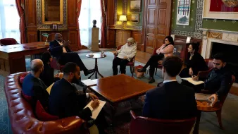 Nell'immagine si vede un incontro formale all'interno di una stanza ben arredata, con pareti in legno e decorazioni artistiche. Un gruppo di persone è riunito attorno a un tavolino centrale. Alcuni partecipanti sono seduti su poltrone, mentre altri sono seduti su uno sgabello o in posizioni più informali. Alcuni hanno in mano blocchi note o penne, suggerendo che stanno prendendo appunti. La luce naturale entra attraverso le finestre, creando un'atmosfera accogliente. Sullo sfondo, si intravedono alcune opere d'arte e un camino, che aggiungono un tocco elegante all'ambiente.