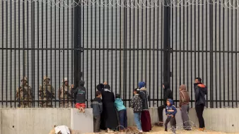 تظهر الصورة مجموعة من الأشخاص الواقفين أمام حواجز حديدية أو سياج مسنن، حيث يواجهون جنود أو عناصر أمنية على الجانب الآخر. يبدو أن هناك أطفالًا ونساء ورجالًا، وبعضهم يحملون أكياسًا أو متعلقات أخرى. البيئة المحيطة موحشة، مع وجود سلك شائك في المقدمة، مما يوضح التوتر والمشاعر القاسية في هذا المشهد.