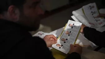 L'image montre une personne tenant un bulletin de vote. En arrière-plan, on peut voir d'autres bulletins similaires. Les bulletins présentent des portraits de candidats avec leurs noms et éventuellement des informations liées à l'élection. L'ambiance semble être celle d'un bureau de vote. La lumière est naturelle et le cadre semble simple, typique d'une situation électorale.