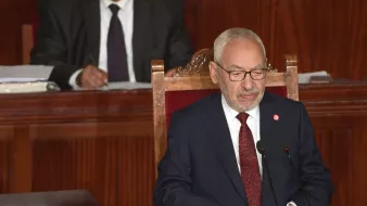 The image shows a man sitting at a podium in a formal setting, likely a legislative or parliamentary assembly. He is wearing a dark suit and glasses, and appears to be speaking or presenting. In the background, another man is seated and observing, dressed in a suit and tie. The environment has a wooden backdrop that contributes to a serious and official atmosphere.