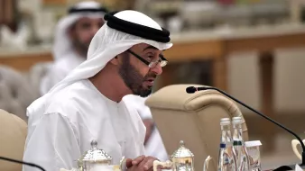 L'image montre un homme portant une tenue traditionnelle émiratie, comprenant une kandura blanche et un ghutrah. Il est assis à une table, apparemment en train de s'adresser à un public. À côté de lui, on peut apercevoir des carafes et des verres d'eau. Le décor semble être celui d'une réunion ou d'une conférence, avec d'autres personnes en arrière-plan. L'atmosphère est sérieuse et formelle.