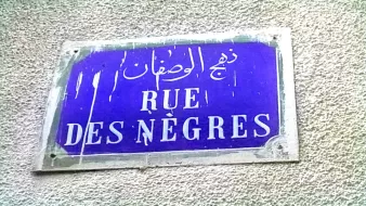 The image shows a street sign. The sign is rectangular and features a blue background with white lettering. It includes text in French that reads "RUE DES NÈGRES," along with Arabic script above it. The sign appears to be weathered, with some signs of wear and tear.