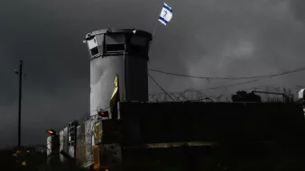 L'image montre une structure semblable à une torre de surveillance, située dans un environnement sombre et nuageux. On peut voir un drapeau israélien flottant au sommet de la tour. Des barbelés sont visibles autour du périmètre, ce qui suggère une zone sécurisée ou protégée. L'éclairage de la scène crée une atmosphère de tension, accentuant l'aspect austère du lieu.