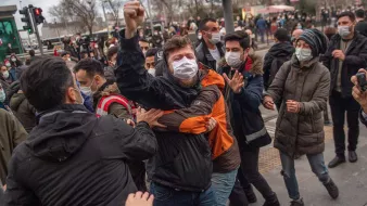 The image depicts a scene of confrontation during a protest or demonstration. Several individuals are gathered, with some appearing to engage in a physical altercation. One person, wearing a black jacket and a mask, is raising a fist, indicating a strong emotional response. People around them seem to be trying to intervene or separate those involved in the conflict. The setting appears to be an urban area, likely near a traffic light, with a backdrop of buildings and other demonstrators. The atmosphere seems tense and charged.
