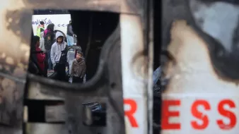 L'image montre un groupe de personnes, y compris des enfants, rassemblées dans un environnement urbain perturbé. On aperçoit une structure endommagée, peut-être un véhicule, qui présente des signes de destruction. Le reflet dans la structure suggère une atmosphère de tension ou de conflit, tandis que les visages des personnes expriment diverses émotions. L'environnement semble chaotique, soulignant une situation difficile.