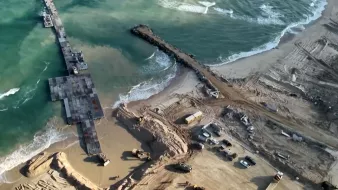 L'immagine mostra una vista aerea di una zona costiera in fase di costruzione o ristrutturazione. Si possono vedere macchine da lavoro e attrezzature presenti sulla spiaggia e nelle vicinanze, mentre un molo si estende verso il mare. Le onde del mare si infrangono sulla riva, e il terreno circostante appare movimentato e scavato, con evidenti segni di lavori in corso. La luce naturale esalta i dettagli del paesaggio, creando un contrasto tra l'acqua blu e la sabbia.