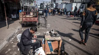 Nell'immagine c'è una strada affollata con diverse persone che si muovono. In primo piano, un uomo è seduto su una sedia accanto a un tavolo, occupato a riparare delle scarpe. Attorno a lui ci sono alcuni oggetti e sacchetti. Sullo sfondo si vedono veicoli e altre persone che camminano. L'atmosfera è vivace e caotica.