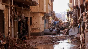 الصورة تظهر مشهداً من منطقة تعرضت لأضرار جسيمة نتيجة كارثة طبيعية، ربما تكون فيضانات أو عواصف. تظهر في الصورة مباني متضررة وجدران متهدمة، وهناك حطام متناثر في الشوارع. كما تُظهر الصورة سيارات مقلوبة أو متضررة، ومياه راكدة تجمعت في الشوارع. المشهد يعكس الأثر المدمر الذي أحدثته الكارثة، مما يدلل على حالة الفوضى والمأساة التي خلفتها.