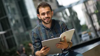 L'image montre un jeune homme assis sur un banc, souriant, tout en tenant un carnet ou un cahier dans ses mains. Il porte une chemise à carreaux et a l'air détendu, avec un environnement urbain en arrière-plan et des bâtiments modernes. La lumière naturelle éclaire la scène, créant une atmosphère agréable.