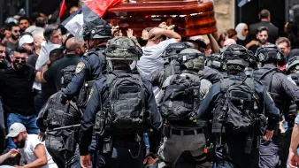تظهر الصورة مشهدًا من حدث عام حيث يحدث تداخل بين مجموعة من الناس وقوات الأمن. يتم حمل تابوت خشبي من قبل المشاركين في الهتافات، بينما يحيط بهم رجال شرطة يرتدون تجهيزات خاصة. الخلفية تضم أعلامًا، مما يشير إلى أن هذه الفعالية قد تكون مرتبطة بمسألة وطنية أو سياسية. الجو يبدو مشحونًا بالانفعالات، مع وجود حشد من الناس في حالة من التوتر.