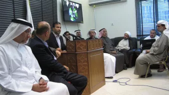L'image montre un groupe de personnes rassemblées dans une salle. Au centre, un homme est en train de parler devant un microphone, tandis que les autres l'écoutent attentivement. Certains d'entre eux portent des vêtements traditionnels, comme des kandouras et des ghutras, indiquant peut-être un contexte culturel spécifique. À l'arrière-plan, un écran affiche probablement une présentation ou une vidéo. L'atmosphère semble sérieuse et concentrée, suggérant que la réunion pourrait porter sur un sujet important.