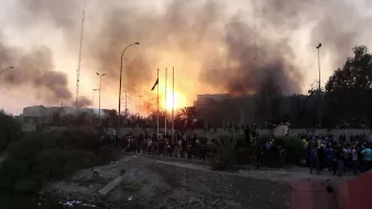 این تصویر نمایی از یک سناریو پرتنش را به تصویر می‌کشد، که شامل آتش و دود در آسمان است. غلظت دود نشان‌دهنده یک حادثه یا اعتراض شدید است. افرادی در پایین تصویر تجمع کرده‌اند و به نظر می‌رسد که در حال تماشا یا شرکت در یک رویداد مهم هستند. در پس‌زمینه، ساختمان‌هایی دیده می‌شود که به همراه تیرک‌های پرچم و تجهیزات دیگر قابل توجه است. این صحنه به طور کلی حس ناامنی و هیجان را منتقل می‌کند.