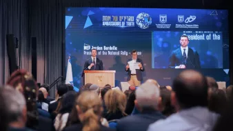 L'image montre une conférence avec un orateur debout derrière un pupitre, lisant un discours. Le public clairement visible est assis devant lui, et un grand écran derrière affiche des informations sur l'événement. L'atmosphère est formelle et engageante.