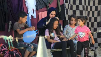 L'immagine mostra un gruppo di bambini e una ragazza seduti all'aperto, in un ambiente semplice. Una donna, vestita con un abito scuro, è intento a pettinare i capelli di un'altra ragazza, che sembra concentrata. Altri bambini sono presenti, alcuni giocano con un palloncino e altri osservano con interesse. Sullo sfondo, si possono vedere vestiti appesi e un ambiente rustico con pietre e teli. L'atmosfera sembra informale e familiare.