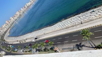 الصورة تظهر مشهداً لجانب من البحر، حيث يمتد الشاطئ على الواجهة البحرية. يحيط بالمكان الطريق الذي يمتد على طول الساحل، ويظهر فيه عدد من الأشجار والنخيل. في الخلفية، يمكن رؤية مجموعة من المباني السكنية أو الفنادق التي تعكس نمط الحياة الحضري. لون المياه يبدو زرقاً صافياً، مما يضيف جمالاً إلى المشهد.