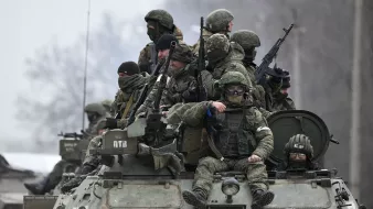 L'immagine mostra un gruppo di soldati equipaggiati con abbigliamento militare e armi, che si trovano su un veicolo blindato. Alcuni di loro indossano maschere o occhiali protettivi, mentre altri appaiono attenti e pronti all'azione. Il contesto è probabilmente un'area di conflitto, con un paesaggio grigio e invernale sullo sfondo. L'atmosfera è seria e tesa, riflettendo la situazione militare.