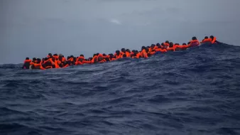 L'image montre un groupe de personnes en mer, portant des gilets de sauvetage orange. Elles semblent rassemblées sur une petite embarcation, luttant contre des vagues dans une étendue d'eau bleue profonde. Le ciel est nuageux, ce qui accentue la gravité de la situation maritime. Cette scène évoque des thèmes de migration, de survie et de danger en mer.