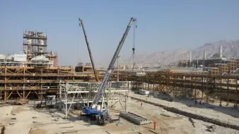 The image shows a large construction site with various structures being built. In the foreground, there is a crane positioned to lift materials, with its arm extended upward. Nearby, there are workers engaged in construction activities. Surrounding the site are several large industrial buildings and infrastructure, indicating that this is likely a major development project. The background features mountains, and the sky is clear. Overall, the scene conveys a busy, active construction environment.