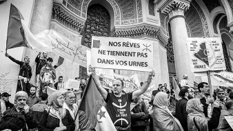 The image depicts a protest scene, where a diverse group of people is gathered. Central to the scene is a man holding a large sign that reads "NOS RÊVES NE TIENNENT PAS DANS VOS URNES," which translates to "Our dreams do not fit in your ballot boxes." The crowd appears to be passionate, with some individuals holding banners, possibly conveying political messages. The setting has an architectural backdrop, suggesting a significant public space, such as a government building or place of gathering. The black and white tones emphasize the seriousness of the demonstration.