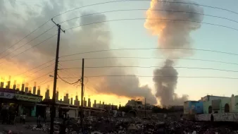الصورة تظهر مشهداً لسماء ملونة بالأصفر والبرتقالي مع وجود سحب من الدخان. يبدو أن هناك انفجارات أو حريق في الخلفية حيث تتصاعد أعمدة من الدخان. في المقدمة، توجد نفايات تتكدس على الأرض مع بعض المباني واللافتات. المشهد يوحي بأجواء متوترة وصعبة.