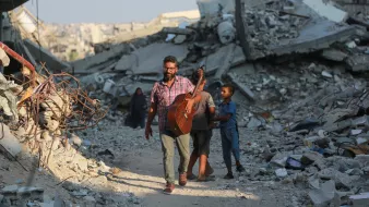 L'immagine mostra un uomo che cammina tra le macerie di un'area distrutta, portando una chitarra. Sembra essere felice mentre si muove, accompagnato da un bambino. Sullo sfondo ci sono molti detriti e edifici in rovina, che suggeriscono una situazione di distruzione. Un'altra figura, probabilmente una donna, è visibile più lontano tra le macerie. L'atmosfera trasmette un contrasto tra la musica e la devastazione circostante.