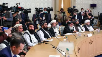L'image montre une réunion importante avec plusieurs hommes assis en rangée derrière une table. Ils portent des vêtements traditionnels et semblent être des membres d'un groupe politique ou de dirigeants. En arrière-plan, il y a de nombreux journalistes et caméras, suggérant que l'événement est médiatisé. La salle est bien éclairée, avec une décoration neutre et un air d'importance formelle.