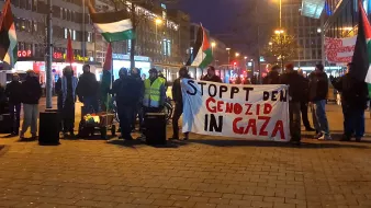 تُظهر الصورة مجموعة من المتظاهرين في مكان عام، يحملون لافتة كبيرة مكتوب عليها "STOPPT DEN GENOZID IN GAZA"، مما يعني "أوقفوا الإبادة الجماعية في غزة" باللغة الألمانية. يُلاحظ في الصورة أيضًا العلم الفلسطيني، حيث يرفع بعض المتظاهرين الأعلام دعمًا للقضية. يبدو أن التظاهرة تُعقد في وقت متأخر من اليوم، حيث تُضاء الأضواء في الشارع المحيط.