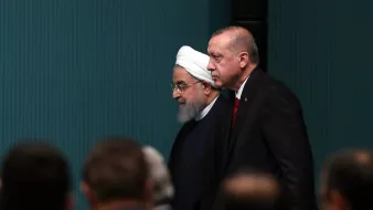 The image shows two men walking side by side. One is wearing a white turban and a black robe, while the other is dressed in a formal suit. They appear to be in a formal setting, likely a conference or meeting, with other individuals partially visible in the foreground. The background features a minimalist wall with a light blue-green color. The atmosphere seems serious and professional.