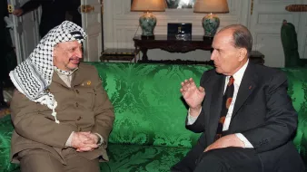 L'image montre deux hommes assis sur un canapé vert décoré. L'un des hommes porte un costume beige avec un keffieh, tandis que l'autre est en costume sombre avec une cravate à motifs. Ils semblent engagés dans une conversation animée. En arrière-plan, on aperçoit des éléments de mobilier et une lumière douce, suggérant un cadre intérieur élégant.