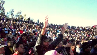 L'image montre une vaste foule de personnes rassemblées dans un espace ouvert. On peut voir des milliers de spectateurs, certains levant les bras en l'air, clairement engagés et enthousiastes. L'arrière-plan est constitué de buissons et d'arbres, tandis que le ciel est bleu et dégagé. L'atmosphère semble festive et dynamique, suggérant un événement important ou un concert.