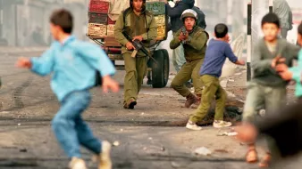L'image montre une scène de tension dans un environnement urbain, où deux soldats sont présents, armés et en position d'action. Un homme, probablement un civil, est également visible, tandis que des enfants et d'autres personnes s'éloignent rapidement de la situation, suggérant un climat de peur ou de chaos. Des véhicules et des baraques sont en arrière-plan, accentuant l'atmosphère de conflit. L'ensemble évoque un contexte de violences ou d'affrontements dans une ville.