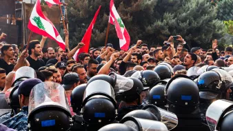 L'image montre une scène de manifestation avec un grand nombre de personnes brandissant des drapeaux libanais. Les manifestants semblent exprimer leurs revendications avec des poings levés, tandis qu'une ligne de policiers en uniforme, équipés de casques et de protections, se tient face à eux. L'ambiance est tense, illustrée par la proximité des manifestants et des forces de l'ordre. Des arbres en arrière-plan complètent le décor de cette confrontation.