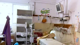 L'immagine mostra una stanza d'ospedale in uno stato di trascuratezza. Si possono notare letti ospedalieri con lenzuola disordinate, attrezzature mediche sparse e un mobile con vari oggetti all'interno. Le pareti sono decorate con disegni infantili, ma l'ambiente appare comunque piuttosto disordinato. Un monitor è appeso al muro e ci sono altri strumenti medici, indicando che si tratta di un'area destinata alla cura dei pazienti. La luce naturale entra dalla finestra, creando un contrasto con l'aspetto poco curato della stanza.
