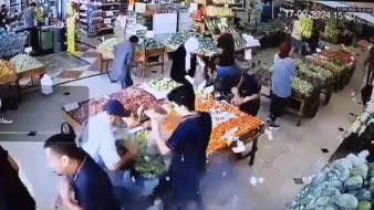 L'image montre une scène animée à l'intérieur d'un marché ou d'une épicerie. On peut voir plusieurs personnes qui interagissent autour de tables chargées de fruits et légumes. L'ambiance semble occupée, avec diverses personnes, certaines portant des sacs, en train de choisir des produits. Les étals sont colorés et bien garnis, suggérant une variété de produits frais disponibles. L'environnement paraît lumineux et accueillant.