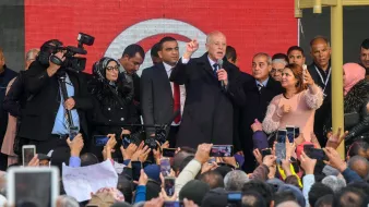 L'image montre un groupe de personnes rassemblées devant une scène où un homme, probablement un leader politique, prend la parole. Il est entouré de plusieurs personnes, dont des membres de la sécurité et des journalistes avec des caméras. La foule est très engagée, beaucoup de gens tiennent des téléphones pour filmer ou prendre des photos. L'arrière-plan présente des affiches ou des bannières, probablement en rapport avec un événement important. L'ambiance semble dynamique et pleine d'énergie.