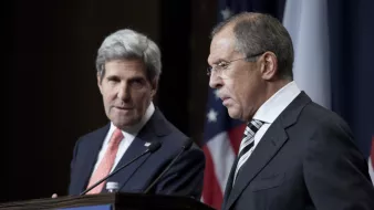 L'image montre deux hommes en tenue formelle, debout à un podium pendant une conférence de presse. L'un est à gauche, avec les cheveux gris et portant un costume bleu, et l'autre à droite, avec les cheveux foncés et portant un costume noir. Ils semblent être en train de discuter sérieusement, avec des drapeaux en arrière-plan, suggérant un contexte diplomatique ou de relations internationales.