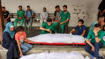 L'image montre un groupe de personnes assises sur des bancs, visiblement en attente dans un espace public. Parmi elles, certains portent des vêtements de santé, tandis que d'autres sont habillés de manière quotidienne. Au premier plan, deux corps enveloppés dans des draps blancs sont visibles, accompagnés de personnes qui semblent éprouver de la tristesse. L'atmosphère générale dégage une forte émotion, suggérant un moment de deuil ou de perte.
