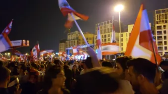 L'image montre une foule rassemblée à la nuit tombée, tenant des drapeaux libanais. Les gens semblent être en plein mouvement, probablement en train de célébrer ou de manifester. On aperçoit une scène à l'arrière-plan, éclairée par des lumières, ce qui suggère un événement festif ou politique. L'atmosphère est dynamique, remplie d'énergie, avec des expressions de joie et d'engagement parmi les participants.