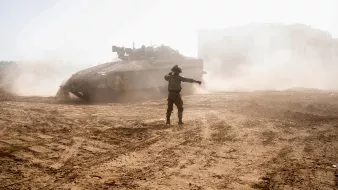 L'immagine mostra un soldato che si trova in un terreno polveroso, mentre un carro armato si muove sullo sfondo. Il soldato sembra indicare qualcosa, con una postura che suggerisce una direzione o un comando. La scena è caratterizzata da una luce intensa, che crea un'atmosfera di tensione, e ci sono nuvole di polvere sollevate dal carro armato, contribuendo a un senso di movimento e caos. Sullo sfondo, si intravede una struttura, ma i dettagli non sono ben definiti a causa della luce e della polvere.