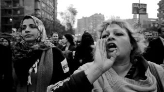 L'image montre une scène de manifestation en noir et blanc. Au premier plan, une femme au cheveux blonds, visiblement passionnée, crie ou exprime une émotion forte, tandis qu'une autre femme, à gauche, porte un foulard et semble attentive. On aperçoit en arrière-plan un grand groupe de personnes, ce qui suggère un mouvement collectif. L'atmosphère est chargée d'énergie et de détermination. Les bâtiments autour ajoutent un contexte urbain à cette démonstration.