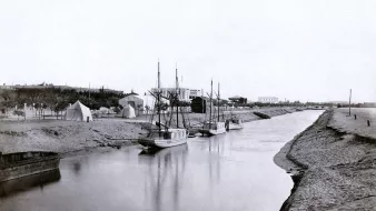 L'image montre une scène tranquille et nostalgique d'un canal ou d'une rivière. On y voit plusieurs bateaux amarrés, leur reflet se mêlant à l'eau calme. En arrière-plan, il y a des tentes et des structures qui évoquent une installation temporaire ou un camp. Le paysage environnant semble naturel, avec de la terre et des arbres. Cette photographie en noir et blanc donne une impression de sérénité et évoque une époque passée.