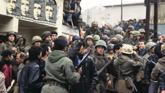 L'image montre un groupe de soldats en uniforme, portant des casques et armés, réunis autour d'une foule. On peut voir des hommes en civil, certains semblent s'opposer aux militaires. En arrière-plan, des affiches sont visibles sur le mur, peut-être représentant des figures politiques ou des messages de protestation. L'atmosphère est tendue et indique un moment de conflit ou de manifestation. Les visages des individus expriment des émotions variées, allant de la détermination à l'inquiétude.