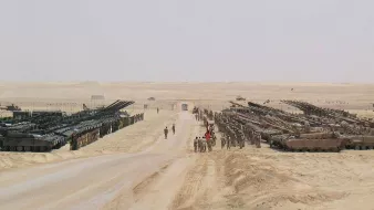 L'image montre un paysage désertique avec des formations militaires. On y voit des chars d'assaut alignés de chaque côté d'une route en terre, tandis que plusieurs soldats sont présents sur le chemin. Le terrain est aride et poussiéreux, avec un ciel clair et peu de végétation. L'atmosphère dégage une impression de préparation militaire ou d'exercice.