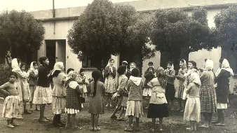 L'image montre un groupe d'enfants, principalement des filles, rassemblés dans une cour. Ils semblent jouer ensemble, entourés de grands arbres. Les filles portent des robes et certaines ont des foulards sur la tête. L'ambiance est joyeuse et festive, suggérant un moment de camaraderie et d'amusement. L'arrière-plan est constitué de murs d'un bâtiment, ce qui donne une atmosphère de communauté.