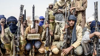 L'image montre un groupe d'hommes armés, probablement dans un environnement désertique. Ils portent des vêtements typiques de régions arides, comme des turbans et des foulards qui cachent partiellement leur visage. Chaque homme tient une arme à feu, et l'ambiance générale semble sérieuse et déterminée. Le groupe est organisé et se présente en ligne, ce qui suggère une certaine discipline. Le fond désertique renforce la dureté du cadre dans lequel ils se trouvent.
