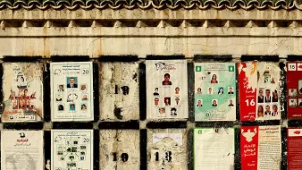 L'image montre un mur sur lequel sont affichés plusieurs documents et affiches. Ces derniers semblent être en lien avec des élections, avec des plaques présentant des photos et des noms de candidats. Certaines affiches sont abîmées et décolorées, révélant un certain vieillissement et une usure du temps. Le mur lui-même présente une texture rugueuse et des motifs décoratifs en haut. Les divers éléments placardés créent une composition visuelle chargée et informative.