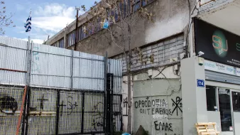 L'image montre un environnement urbain avec un bâtiment en arrière-plan et un mur métallique en façade, où l'on peut voir des graffitis. Un drapeau grec est visible, ainsi qu'un espace de stationnement ou de stockage à côté d'une entrée. Le sol semble en béton, et quelques déchets sont éparpillés sur le sol. Les graffitis sur le mur comprennent des inscriptions en grec, ce qui suggère un contexte local ou culturel. L'atmosphère paraît un peu désordonnée, typique des zones industrielles ou abandonnées.
