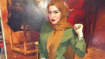 L'immagine mostra una giovane donna che indossa un hijab color marrone e una camicia verde. È in piedi con una mano sulla vita e l'altra sollevata, con un'espressione sicura. Indossa jeans attillati e scarpe marroni. Sullo sfondo si intravede un dipinto o una decorazione murale che aggiunge un tocco di colore all'ambiente. L'illuminazione sembra calda, contribuendo a un'atmosfera accogliente.
