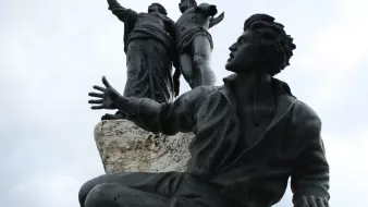 L'image montre une sculpture en bronze représentant plusieurs figures humaines. Au premier plan, un homme est assis, les bras ouverts, avec une expression qui semble transmettre une émotion forte. Derrière lui, deux figures debout sont également visibles, levant les bras, ce qui pourrait suggérer un message de célébration ou de libération. La sculpture est placée sur une base rocheuse, ajoutant une dimension texturisée à l'ensemble. Le ciel apparaît nuageux, ce qui donne un ton dramatique à la scène.